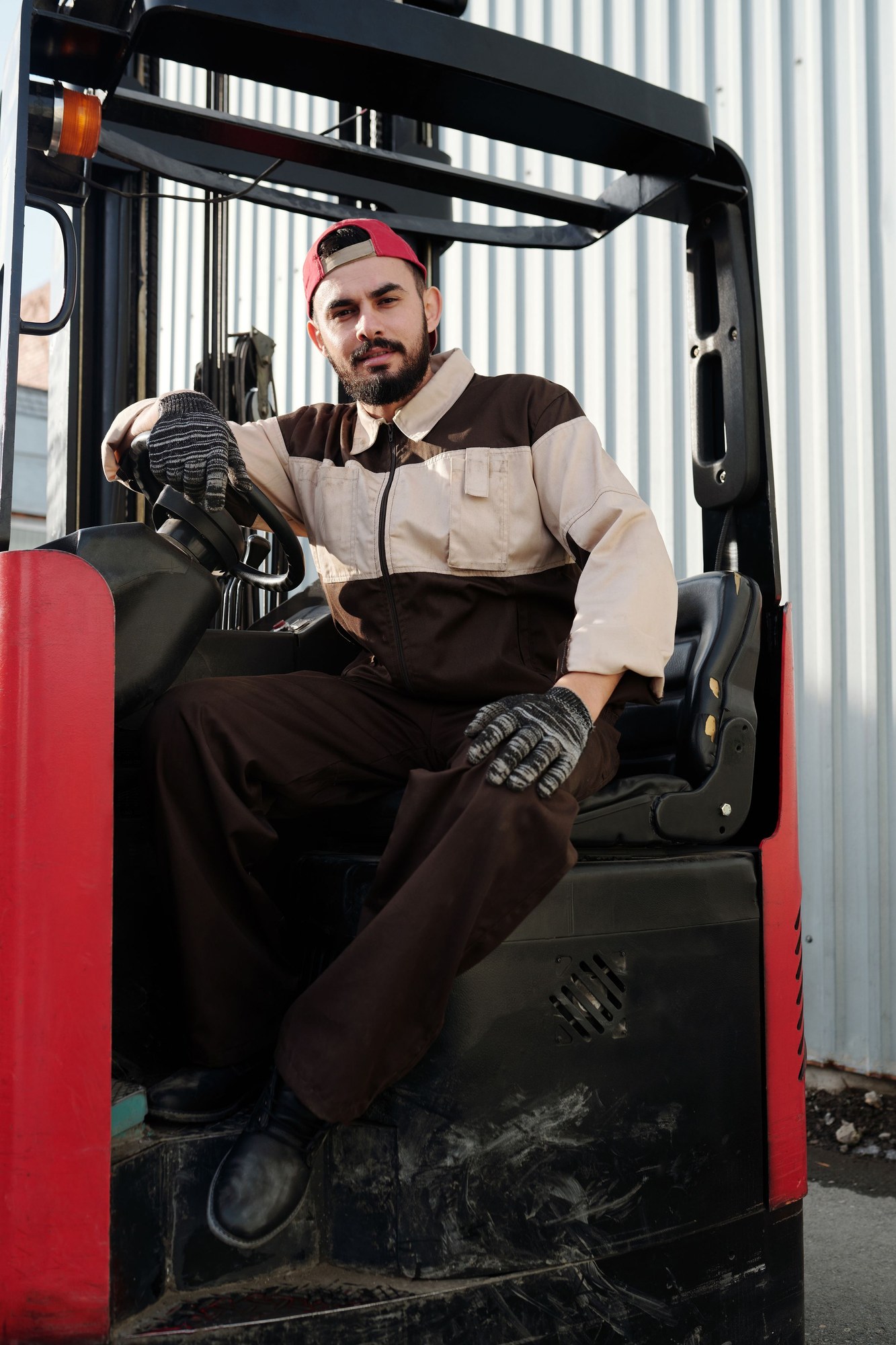 A warehouse operator wearing work gloves sits at the controls of a red forklift.